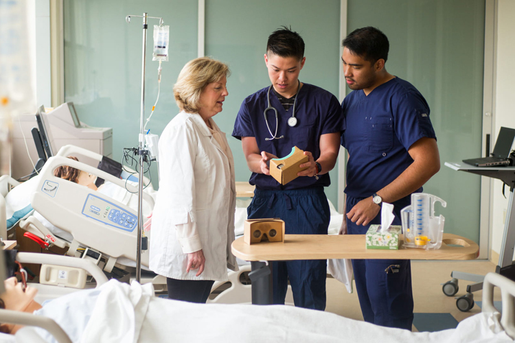 health students in scrubs look at clipboard with professor in lab coat.  Clinic setting with dummy in hospital bed in foreground.