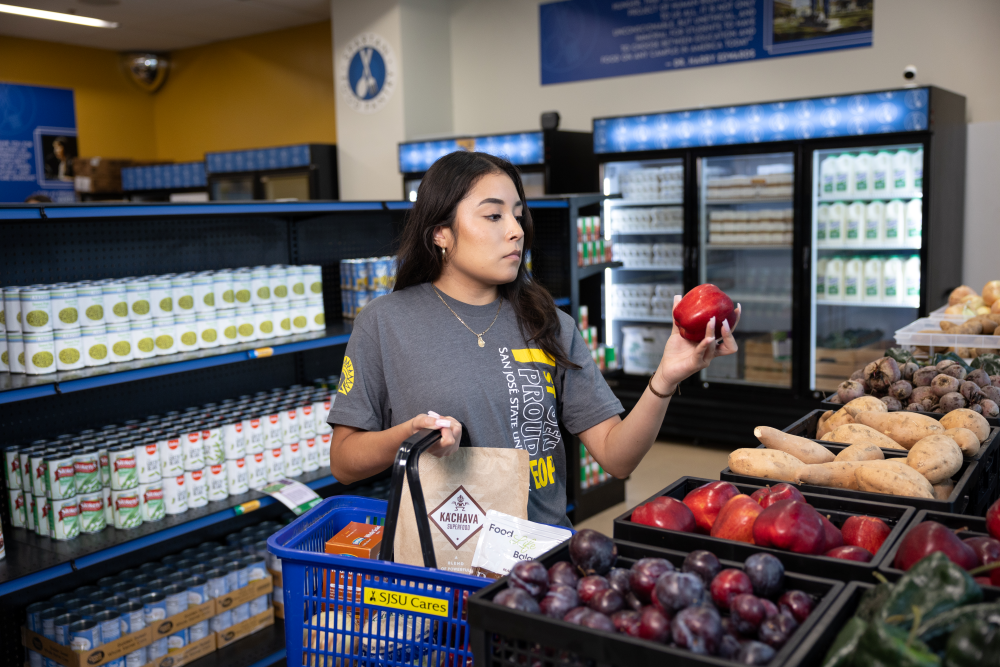 A student shops at the SJSU Spartan Food Pantry, holding a red apple and a basket filled with groceries, surrounded by fresh produce and pantry staples.