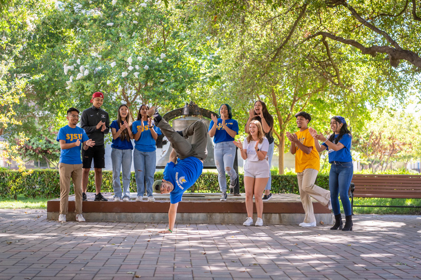 Students hanging out and dancing on campus. Greenery surrounds them with tower bell right behind them.